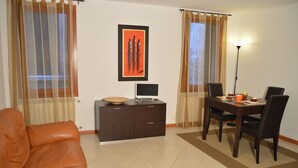 Un salon avec un canapé marron, un meuble en bois foncé sur lequel se trouve un ordinateur portable, un tableau encadré accroché au mur, et un coin repas avec une table et des chaises.