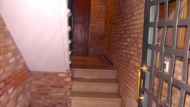 Un escalier dont les marches sont recouvertes d’un carrelage à motifs en damier, menant à une porte ornée d’un motif en treillis métallique.