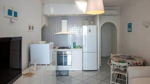 A compact kitchen with a refrigerator, oven, and sink, a dining table with a floral tablecloth, and a TV on the left.