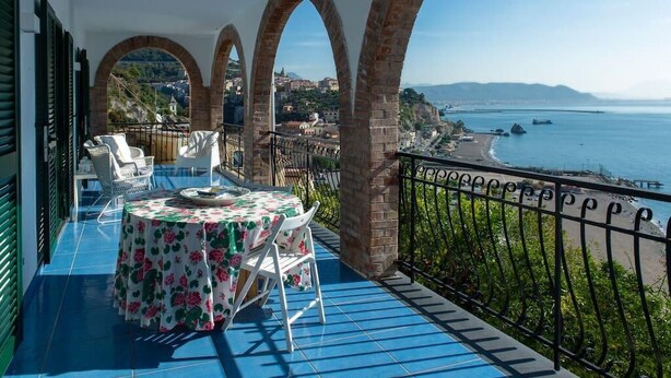A balcony with a table and chairs, overlooking a coastal town and the sea.