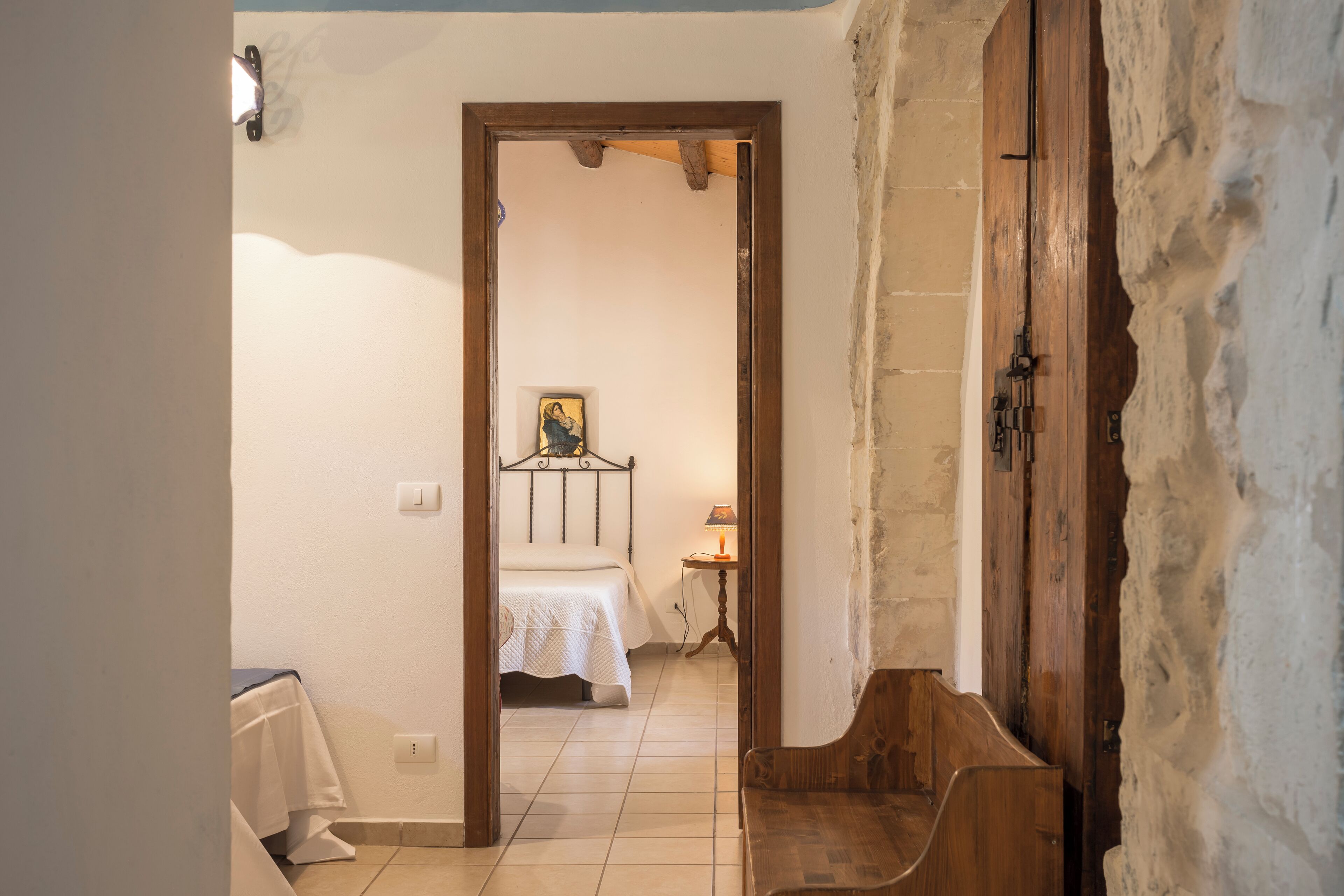 Un couloir étroit mène à une chambre à coucher qui contient un lit, une table de chevet et une lampe. Un banc en bois se trouve dans le couloir.