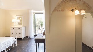 Ein Schlafzimmer mit Bett, Schminktisch und Nachttisch. Ein Fenster mit Vorhängen, eine Tür zu einem Balkon und eine Wandlampe sind vorhanden.