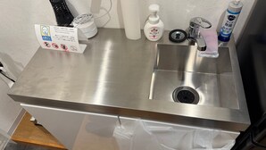 A stainless steel sink with a plastic bag hanging below it, a paper towel dispenser, a bottle of hand soap, and a sign with a no-smoking symbol.