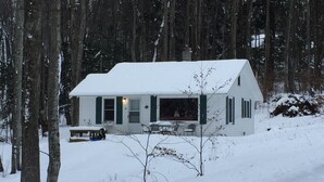 A white cabin with a porch, surrounded by trees and covered in snow.