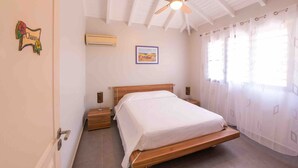 A bedroom with a wooden bed, a ceiling fan, a nightstand, a wall-mounted air conditioner, and a window with sheer curtains. - Treat yourself to a dream vacation! (Petit Bourg)
