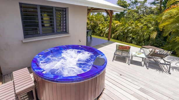 A hot tub on a wooden deck with a covered patio area, a window with blinds, and outdoor furniture. - Treat yourself to a dream vacation! (Petit Bourg)