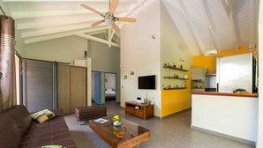A spacious living area with a brown leather sofa, a glass coffee table, a ceiling fan, and a kitchen area with a white refrigerator and yellow walls. - Treat yourself to a dream vacation! (Petit Bourg)