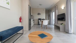 Ein modernes Wohnzimmer mit einer blauen Bank, einem Holztisch mit einem Buch, einer Küchenzeile mit Esstisch und Stühlen sowie einem an der Wand befestigten Fernseher.