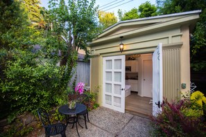 A small, enclosed patio area with a table and chairs, surrounded by lush greenery and a door leading to a kitchen. - Cozy cottage in upscale Claremont Court (Berkeley)