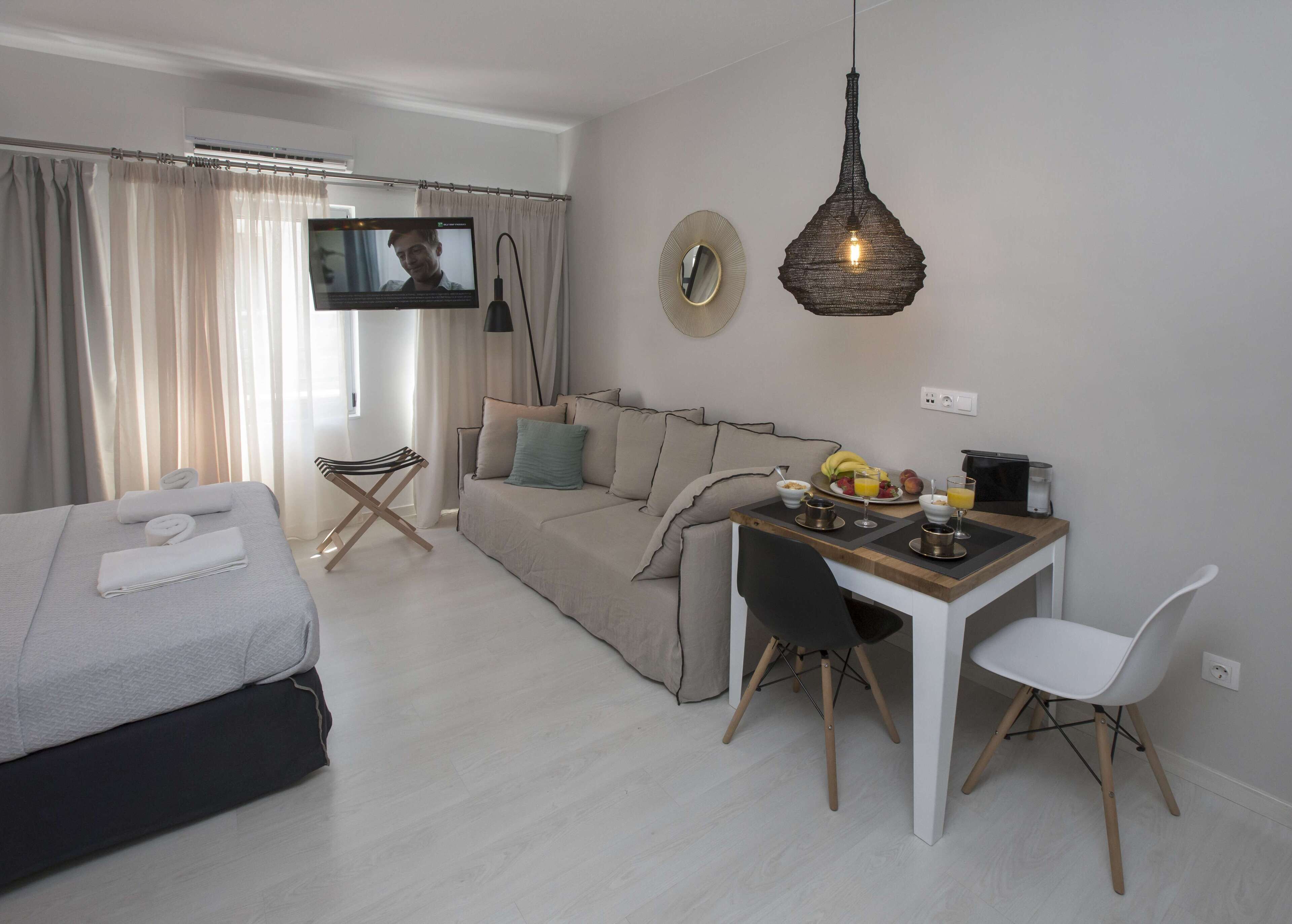 A modern living room with a sofa, a dining table with chairs, a TV mounted on the wall, and a hanging light fixture.
