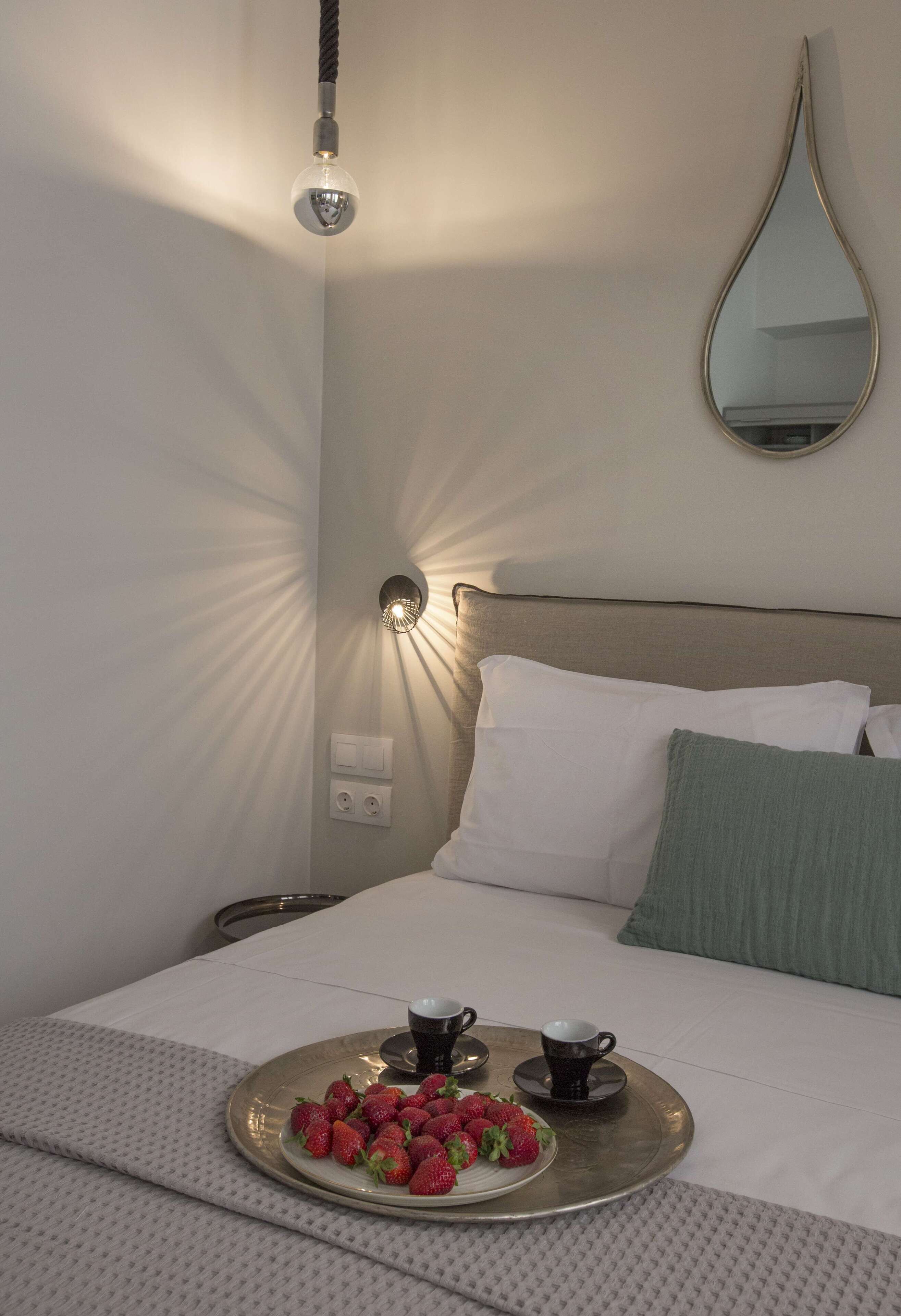 A neatly made bed with a tray of strawberries and two cups, a wall-mounted light, and a mirror.