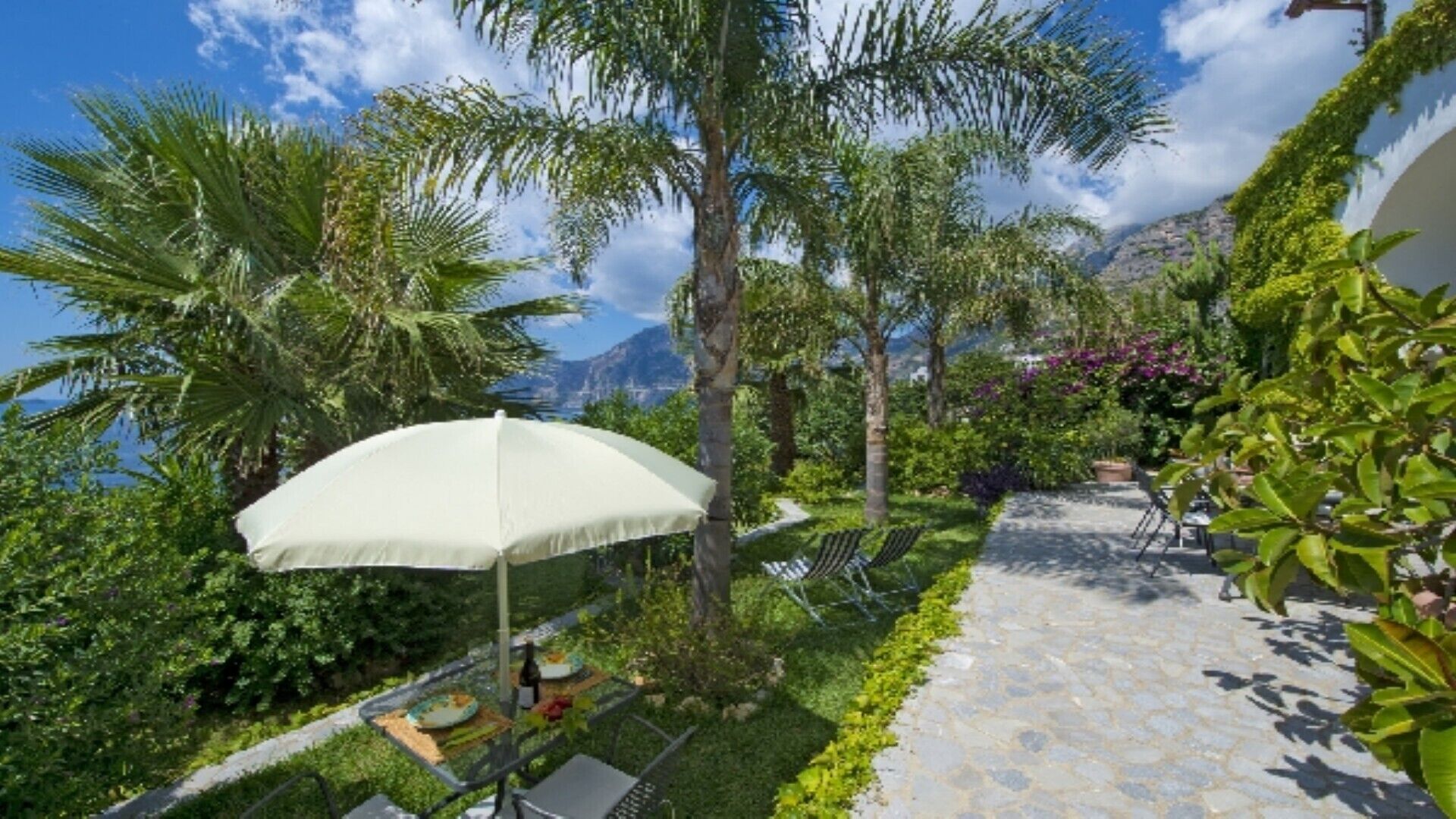 A garden patio with a white umbrella, a table with chairs, and a view of mountains.