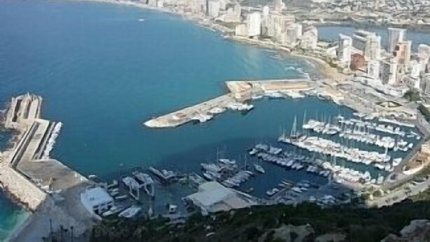 Paesaggio costiero con un porto turistico, edifici e montagne sullo sfondo.
