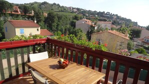 A wooden table with a basket of fruit on a balcony with red railings, overlooking a hillside with buildings. - Collioure: beautiful apartment classified ***, quiet, swimming pool, garden, private parking (Collioure)