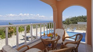 Ein Balkon mit Korbmöbeln, einem Tisch mit Früchten und einem Glas, sowie Blick auf einen Pool und Berge.