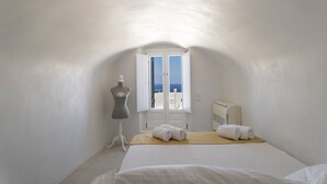 A small, white-walled bedroom with a bed, a mannequin, and a window with a view of the sea. - Oia Mansion 4 Bedroom Villa With Panoramic Sunset View (Santorini)