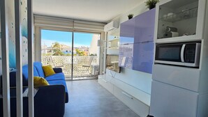 A modern kitchen with a built-in microwave and cabinets, a blue sofa with yellow cushions, and a sliding glass door leading to a balcony with a view of buildings. - Nice - Sapphire studio 40 meters from the sea, 5 p. elevator AC/Wifi, balcony. (Nice)