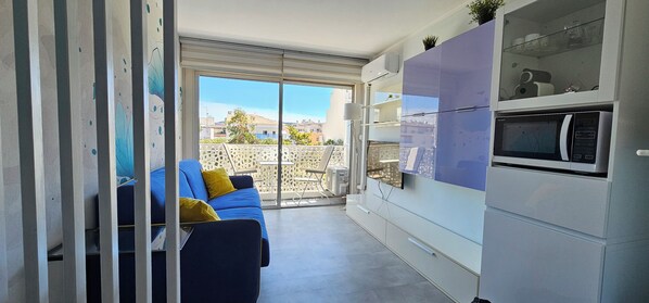 A modern kitchen with a built-in microwave and cabinets, a blue sofa with yellow cushions, and a sliding glass door leading to a balcony with a view of buildings.