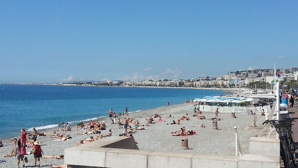 A beach with people sunbathing and swimming, a clear blue sky, and buildings in the background.