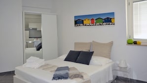 A neatly made bed with white linens and two pillows, a painting of colorful houses on the wall, and a window with blinds. - App. PORTO RUSSO♡ Splendid studio apartment (Otranto)