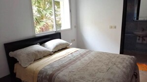 A bedroom with a bed, a window with a view of palm trees, and a bathroom visible through an open door. - Beautiful apartment in Essaouira 4263 (Essaouira)