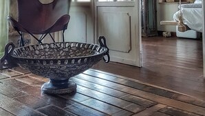 A room with a wooden floor, a metal bowl on a table, a chair, and a bed with a white cover. - Luxurious Stone House in Elani (ΑΜΑ : 00000239911) (Siviri, Chalkidiki)