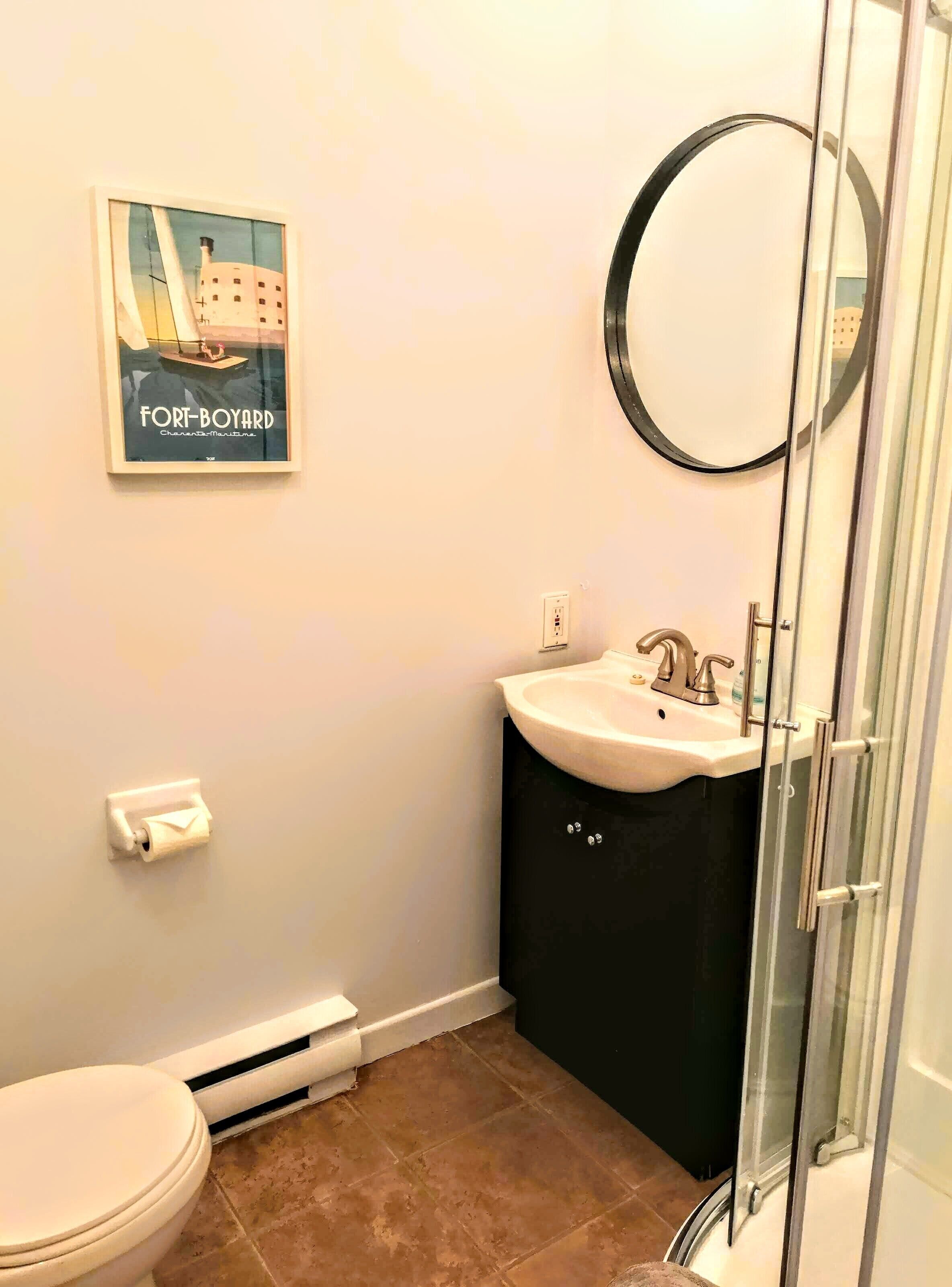 A bathroom with a toilet, a sink, and a shower. There is a framed picture on the wall and a mirror above the sink.
