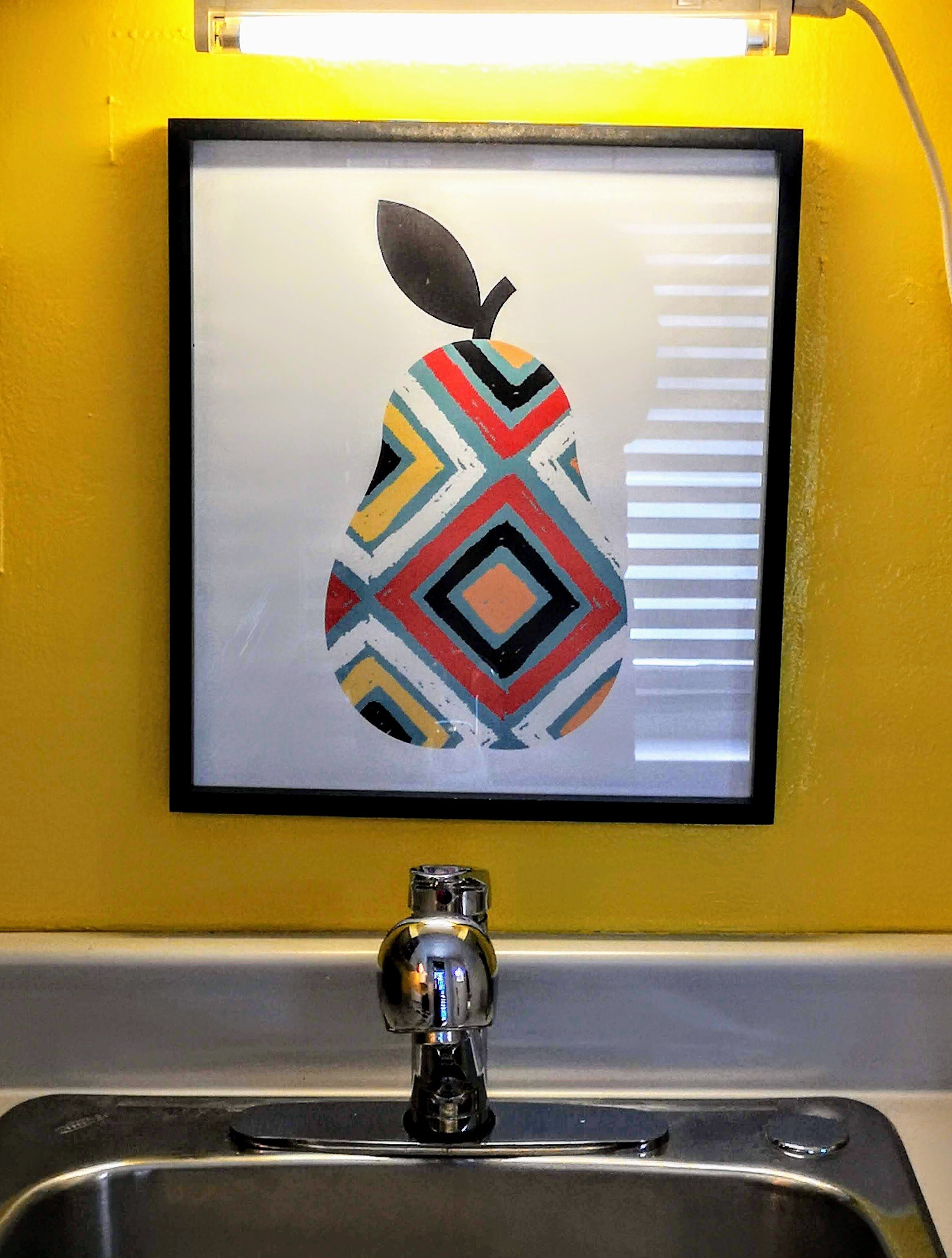 A framed artwork of a stylized pear with a leaf on the wall above a sink.