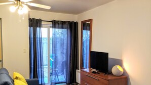 Uma sala de estar moderna com um sofá azul escuro, um armário de madeira com uma televisão de tela plana, um ventilador de teto e uma vista através de uma porta de vidro.