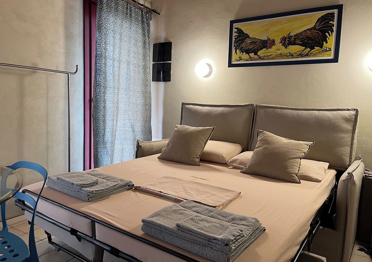 Una cama bien tendida con ropa de cama y almohadas color beige, una mesita de noche con toallas dobladas y una lámpara fijada a la pared.