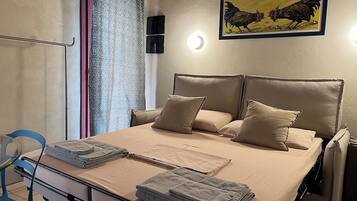 Una cama bien tendida con ropa de cama y almohadas color beige, una mesita de noche con toallas dobladas y una lámpara fijada a la pared.