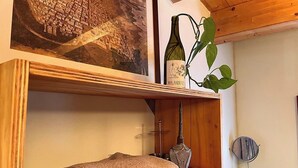 Quarto com teto de madeira, pintura emoldurada de paisagem urbana, garrafa de vinho Malandrin, planta, toalha dobrada, objeto de metal decorativo e um pequeno espelho redondo.