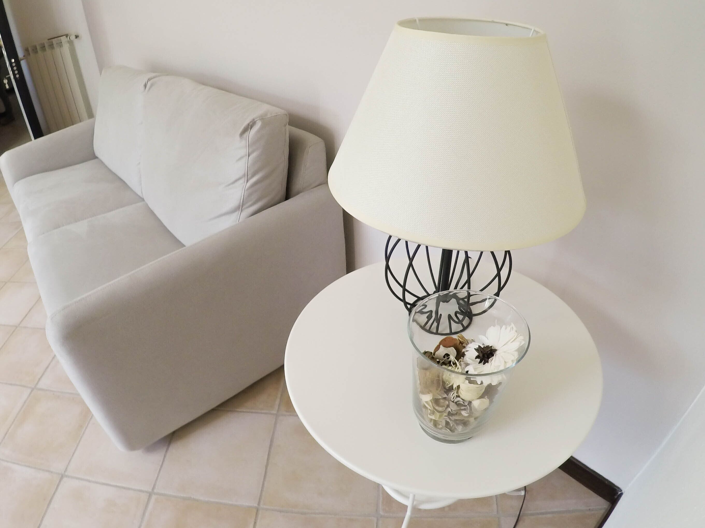 A white sofa with a cushion, a white table with a lamp, and a glass vase with dried flowers.