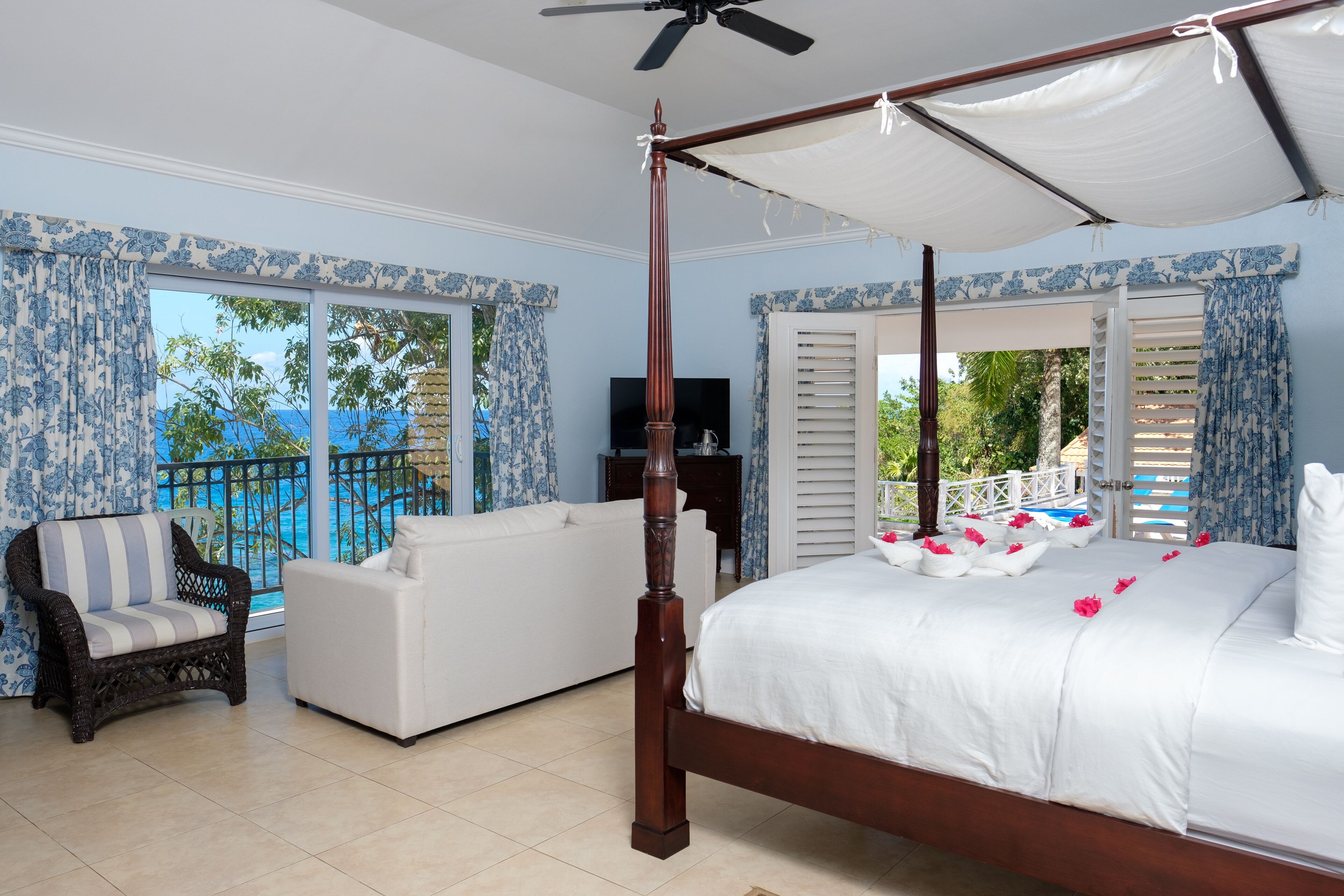 A bedroom with a four-poster bed, a sofa, a chair, and a balcony with a view of the sea.