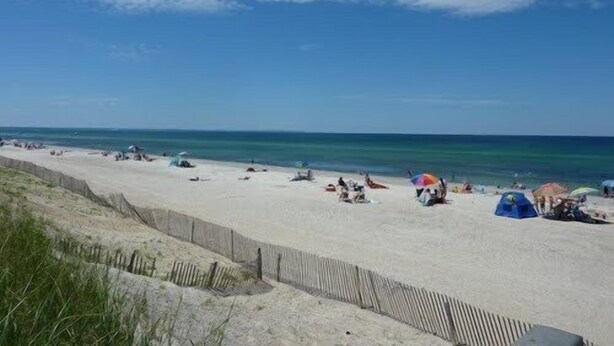 Ein Sandstrand mit Zelten und Sonnenschirmen, eine Holzlattenzäunung und das Meer im Hintergrund.