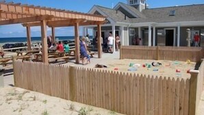 Ein Küstenbereich mit Holzgebäuden, einer überdachten Sitzgelegenheit und einem Sandspielbereich mit Spielzeug.