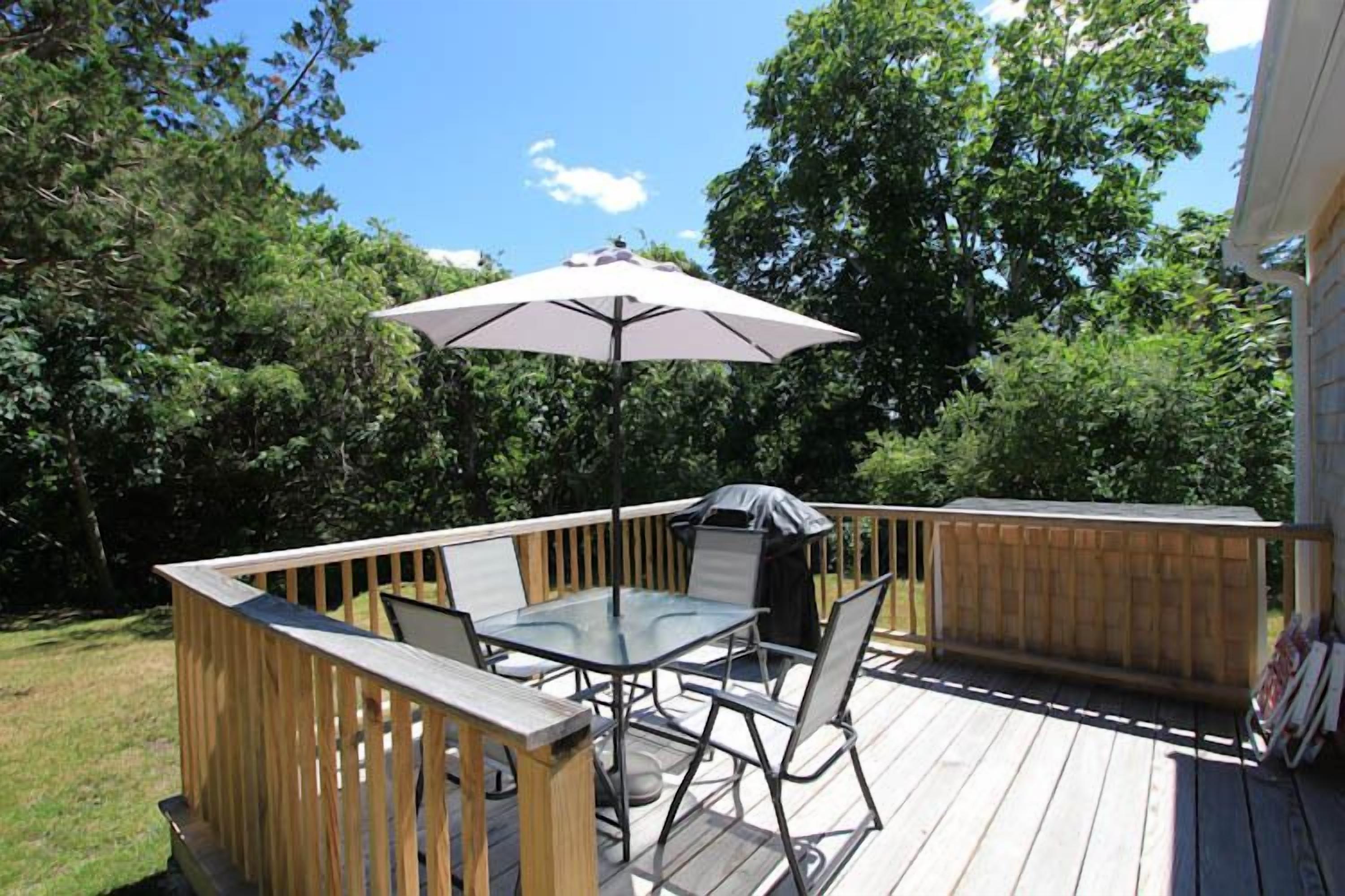 A wooden deck with a glass table and chairs, an umbrella, and a barbecue grill.