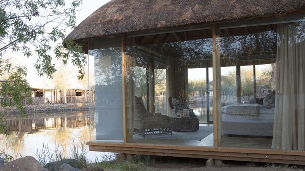 A glass-enclosed room with a thatched roof, a bed, and a wicker chair by a body of water. - Norotshama River Resort (Aussenkehr)