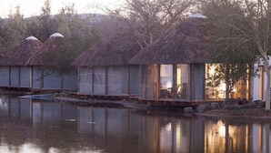 A row of thatched-roof cabins with glass windows, situated by a calm lake, reflecting the structures and trees. - Norotshama River Resort (Aussenkehr)