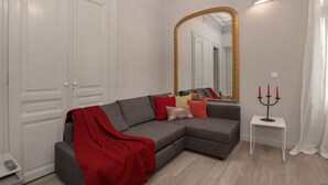 Un salon avec un canapé d’angle gris, un plaid rouge, une petite table d’appoint blanche surmontée d’un chandelier, et un grand miroir avec un cadre doré.