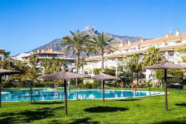 Relax by the resort's large pool surrounded by lush greenery and palm trees, with a mountain view in the background.