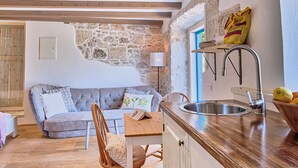 Eine gemütliche Küche mit Steinwand, Holzbalken, einem grauen Sofa, einem Holzessttisch und einem Spülbecken mit Obstschale.
