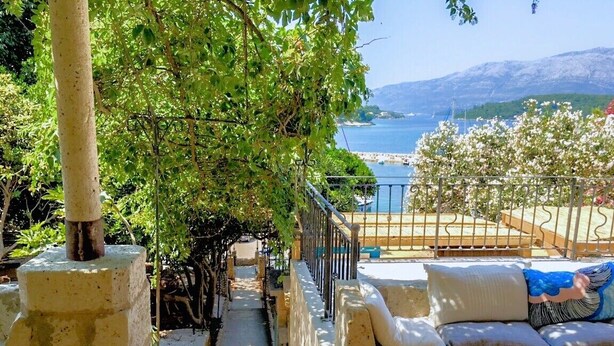 Eine Terrasse mit Meerblick, eine Steinsäule, ein weißer Vasen und eine Couch mit Kissen.
