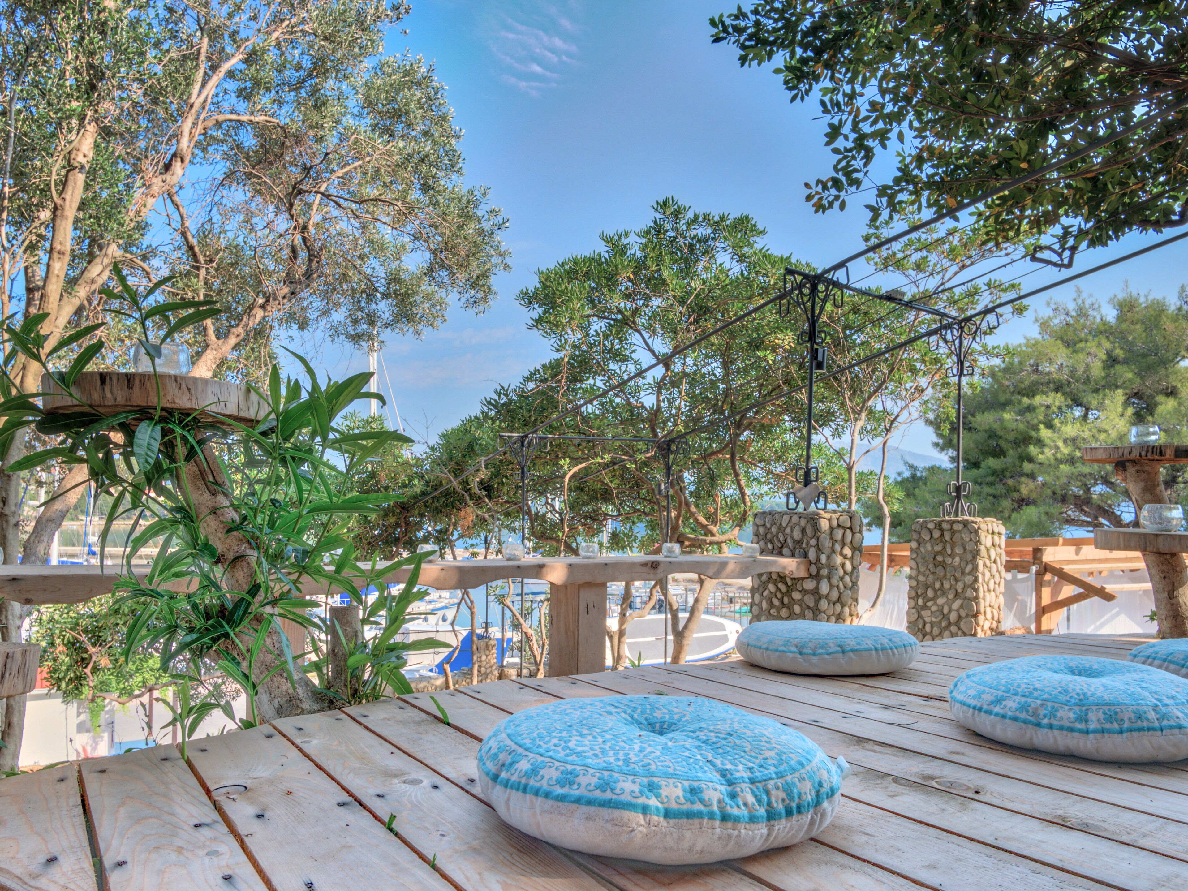 Eine Holzterrasse mit runden Kissen, einer Steinsäule und Blick auf Boote und Bäume.