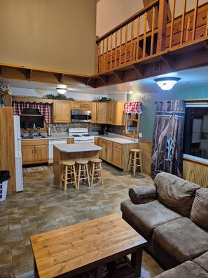 Una cocina con armarios de madera, una isla central con taburetes, una mesa de comedor y un sofá en la sala de estar.