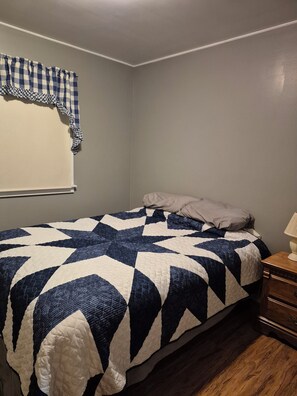 Un dormitorio con cama, mesita de noche, lámpara, ventana con cortina a cuadros y un zócalo en la pared.