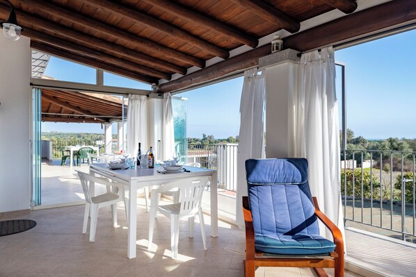 A balcony with a dining table set for two, a blue cushioned chair, and a view of the landscape.