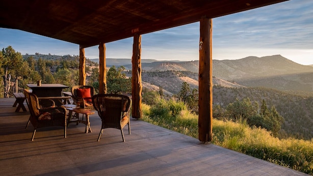 An outdoor deck with wooden pillars, wicker furniture, and a view of a mountainous landscape.