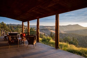 Una terraza al aire libre con pilares de madera, muebles de mimbre y vistas a un paisaje montañoso.