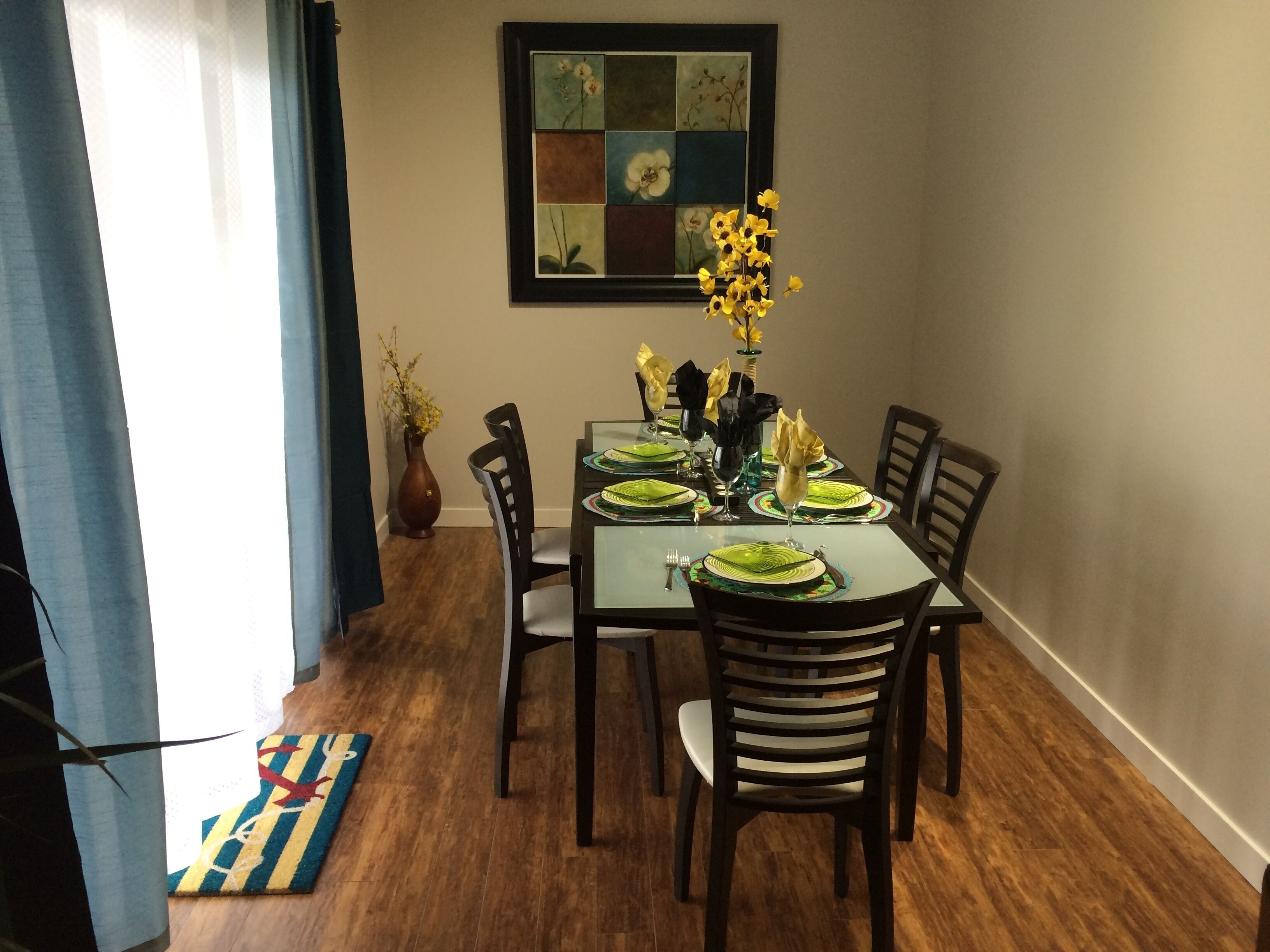 A dining area with a table set for six, chairs, a vase with yellow flowers, and a framed artwork on the wall.